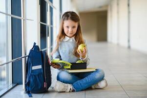 frente ver de pequeño hermosa colegio niña entre corredor a escuela, participación notas a manos. gracioso y contento niña sonriente a cámara, descansando después lecciones en primario colegio foto