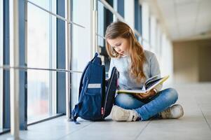 Front view of pretty blonde school girl holding many colorful notes and books. Clever teen girl smiling at camera, standing on corridor of international school. photo