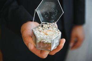 The groom on his wedding day holds two wedding rings in his hand in a hotel room. The man is wearing a white shirt and blue trousers with a vest. Male hand and wedding rings close up. photo