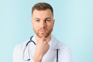 Portrait young bearded doctor with stethoscope over neck in medical coat standing against isolated blue background. photo