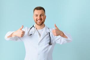 Waist up portrait view of the smiling happy doctor showing big finger at the free space on blue background. Doctor and medicine concept photo