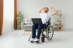 Portrait of smiling disabled male sitting in wheelchair and working on laptop from home. Young worker with special needs. Freelancer and people with disabilities concept. photo