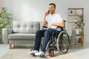 The disabled person sits in a wheelchair. He is talking to someone on his smartphone. He is in his large bright living room. He smiles. photo