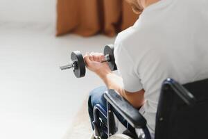 sportsman sitting in wheelchair and outstretching arms with dumbbells during rehabilitation exercise in modern medical center. photo