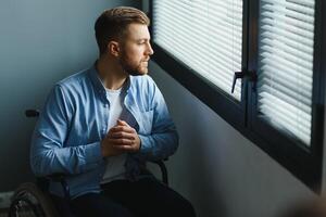 Disabled Person Sits in Wheelchair Against Window. Serious Sad Caucasian Man Wearing Casual Clothes and Look at Large Panoramic View in Bright Modern Living Room or Hospital. photo