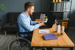 Disabled person in the wheelchair works in the office at the computer. He is smiling and passionate about the workflow photo