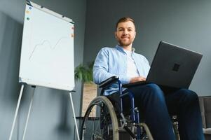 Young man with special needs in casual clothes working on wireless laptop. Male freelancer working from home while sitting in wheelchair. photo