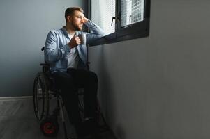 Disabled Person Sits in Wheelchair Against Window. Serious Sad Caucasian Man Wearing Casual Clothes and Look at Large Panoramic View in Bright Modern Living Room or Hospital photo