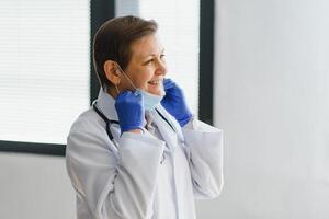 más viejo hembra médico vistiendo cara máscara y blanco médico Saco en pie en hospital. retrato foto