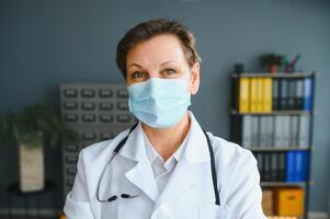 más viejo hembra médico vistiendo cara máscara y blanco médico Saco en pie en hospital. retrato foto