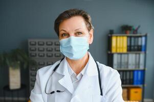 Older female doctor wearing face mask and white medical coat standing in hospital. Portrait photo