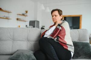 Middle-aged 50s sick frozen woman seated on sofa in living room covered with warm plaid sneezing, runny nose feels unhealthy, seasonal cold, weakened immune system concept photo