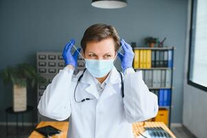 Older female doctor wearing face mask and white medical coat standing in hospital. Portrait photo