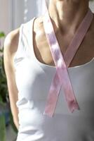 Shot of the woman in the white top against the white wall, with pink ribbon on her neck as a symbol of breast cancer awareness. Concept photo