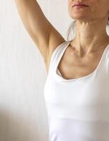 Shot of the woman in the white top against the white wall, performing self examination of the breasts, looking for abnormalities. Breast cancer awareness photo