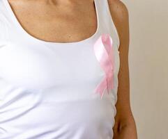 Shot of the woman against the white wall in the white top with pink ribbon, as a symbol of a breast cancer awareness. Concept photo