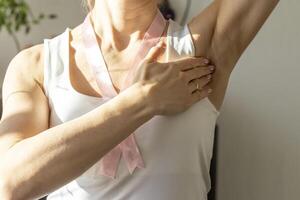 Disparo de el mujer en el blanco arriba, con rosado cinta en su cuello como un símbolo de pecho cáncer conciencia, ejecutando yo examen de el pechos, mirando para anormalidades. concepto foto