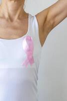 Shot of the woman against the white wall in the white top with pink ribbon, as a symbol of a breast cancer awareness. Concept photo