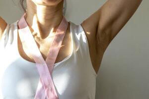 Shot of the woman in the white top against the white wall, with pink ribbon on her neck as a symbol of breast cancer awareness. Concept photo