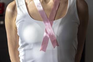 Shot of the woman in the white top against the white wall, with pink ribbon on her neck as a symbol of breast cancer awareness. Concept photo