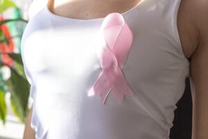 Shot of the woman against the white wall in the white top with pink ribbon, as a symbol of a breast cancer awareness. Concept photo