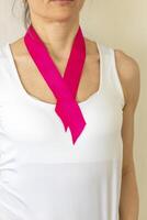 Shot of the woman in the white top against the white wall, with pink ribbon on her neck as a symbol of breast cancer awareness. Concept photo