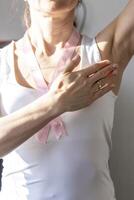 Shot of the woman in the white top, with pink ribbon on her neck as a symbol of breast cancer awareness, performing self examination of the breasts, looking for abnormalities. Concept photo
