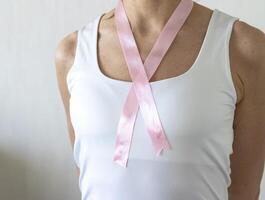 Shot of the woman in the white top against the white wall, with pink ribbon on her neck as a symbol of breast cancer awareness. Concept photo