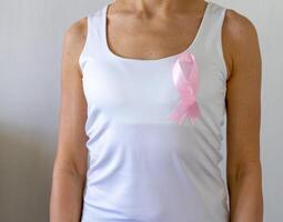 Shot of the woman against the white wall in the white top with pink ribbon, as a symbol of a breast cancer awareness. Concept photo