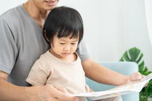 contento asiático padre relajarse y leer libro con bebé hora juntos a hogar. padre sentar en sofá con hija y leyendo un historia. aprender desarrollo, cuidado de niños, reír, educación, contar historias, práctica. foto