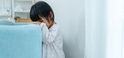 little asian children kid playing hide and seek standing behind the sofa, Adorable child having fun in the home. happy family have fun with kids, activity, learning, activity, game, meditation photo