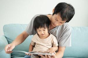 contento asiático padre relajarse y leer libro con bebé hora juntos a hogar. padre sentar en sofá con hija y leyendo un historia. aprender desarrollo, cuidado de niños, reír, educación, contar historias, práctica. foto