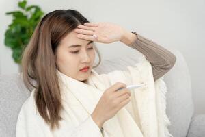 joven asiático mujer teniendo alto fiebre mientras comprobación cuerpo temperatura, hembra estornudos y líquido nariz con estacional influenza, alérgico, digital termómetro, virus, coronavirus, enfermedad, respiratorio foto