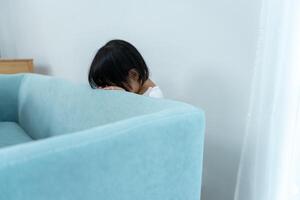 little asian children kid playing hide and seek standing behind the sofa, Adorable child having fun in the home. happy family have fun with kids, activity, learning, activity, game, meditatio photo