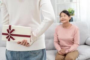 adolescente muchachas son ocultación regalo cajas para su hermosa mamás detrás su espaldas mientras mamá es sentado en el sofá a hogar. celebrando especial ocasión juntos a hogar, contento cumpleaños a mamá foto