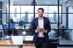 Portrait of Caucasian business man CEO is standing in the office at table with digital tablet and showing statistic chart showing annual report and skyscraper background photo