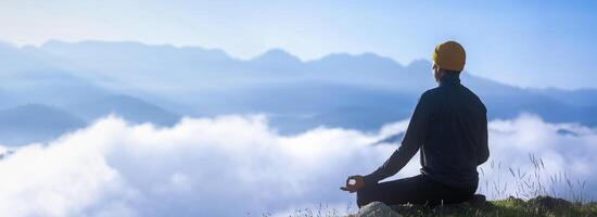 Panorama back view of man is relaxingly practicing meditation yoga mudra at mountain top with mist and fog in summer to attain happiness from inner peace wisdom for healthy mind and soul concept photo