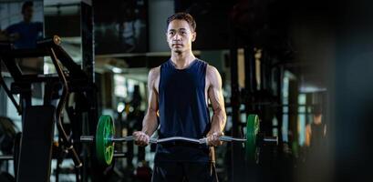 Asian muscular man is practice weight lifing using easy bar as beginner on barbell for arm and core muscle inside gym with dark background for exercising and workout photo