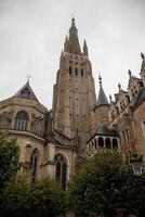 el imponente aguja de el Iglesia de nuestra dama en brujas en contra un nublado cielo - medieval Enladrillado maravilla foto