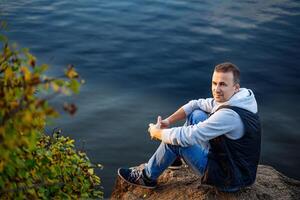 Man Finding Solace on a Rocky Outcrop Overlooking Serene Waters. A man sitting on top of a rock next to a body of water photo