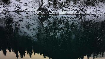 aéreo. reflexión de cubierto de nieve abeto arboles en el superficie de un montaña lago video