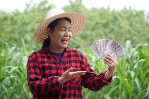 Happy Asian woman wears hat, red plaid shirt, holds Thai banknote money at garden.Concept, agriculture occupation. Thai farmer lifestyle. Satisfied to get income, profit or supporting money. photo