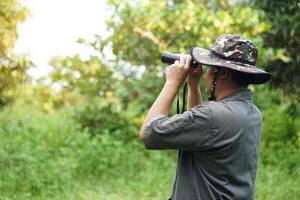 Asian man ecologist use binocular to survey nature in forest. Concept, nature exploration. Ecology study. Pastime activity, lifestyle. Explore environment. Traveling alone photo