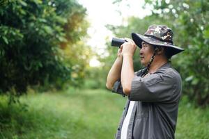 Asian man ecologist use binocular to survey nature in forest. Concept, nature exploration. Ecology study. Pastime activity, lifestyle. Explore environment. Traveling alone photo