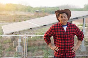 Handsome Asian man wears hat, red plaid shirt, stand beside solar panel at agriculture land, put hands on waists. feels confident. Concept, farmer installed photovoltaic for use in agriculture. photo