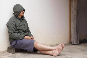 Asian man wears hoodie shirt, shorts, bare feet, sits lean old wall on pavement. Concept, Homeless person. Beggar. Poor. Spy man in unidentified man. photo