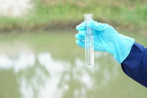 Closeup hand wears blue glove holds test tube of sample water from lake. Concept, explore, inspect quality of water from natural source, prepare for doing experiment. Test of water pollution. photo