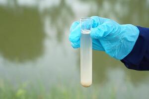 Closeup hand wears blue glove holds test tube of sample water from lake. Concept, explore, inspect quality of water from natural source, prepare for doing experiment. Test of water pollution. photo