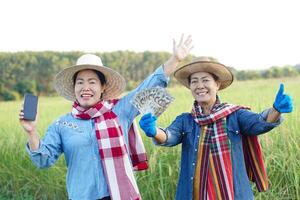 Happy Asian women farmers are at paddy field. They hold smart phone and Thai banknotes money. Concept, Agriculture occupation. Thai farmers. Profit, income. Online apps for promote products photo