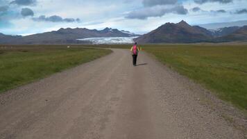 Female tourist with backpack goes by dirt road on background of mountains and glacier in Iceland. Freedom and travel concept. 4K video